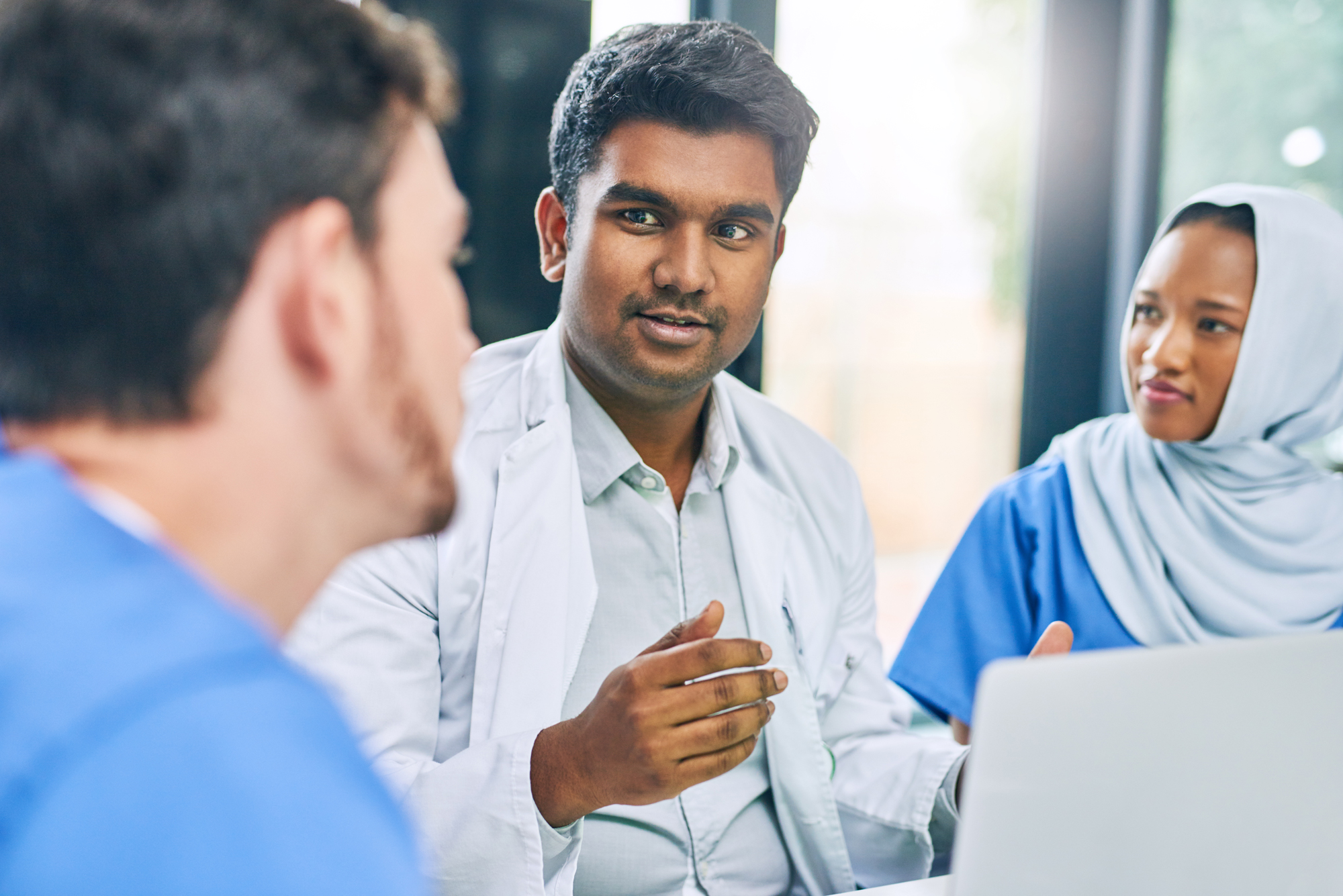 Male doctor in white coat speaking with female patient wearing headscarf and another person, representing patient-centered healthcare communication
