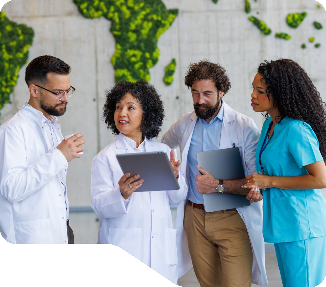 Four healthcare professionals in medical uniforms collaborating with tablets and documents in a modern clinical setting with green plant background